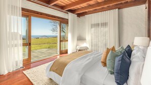 A bedroom with a large bed, wooden ceiling beams, a view through glass doors, and a white lamp.