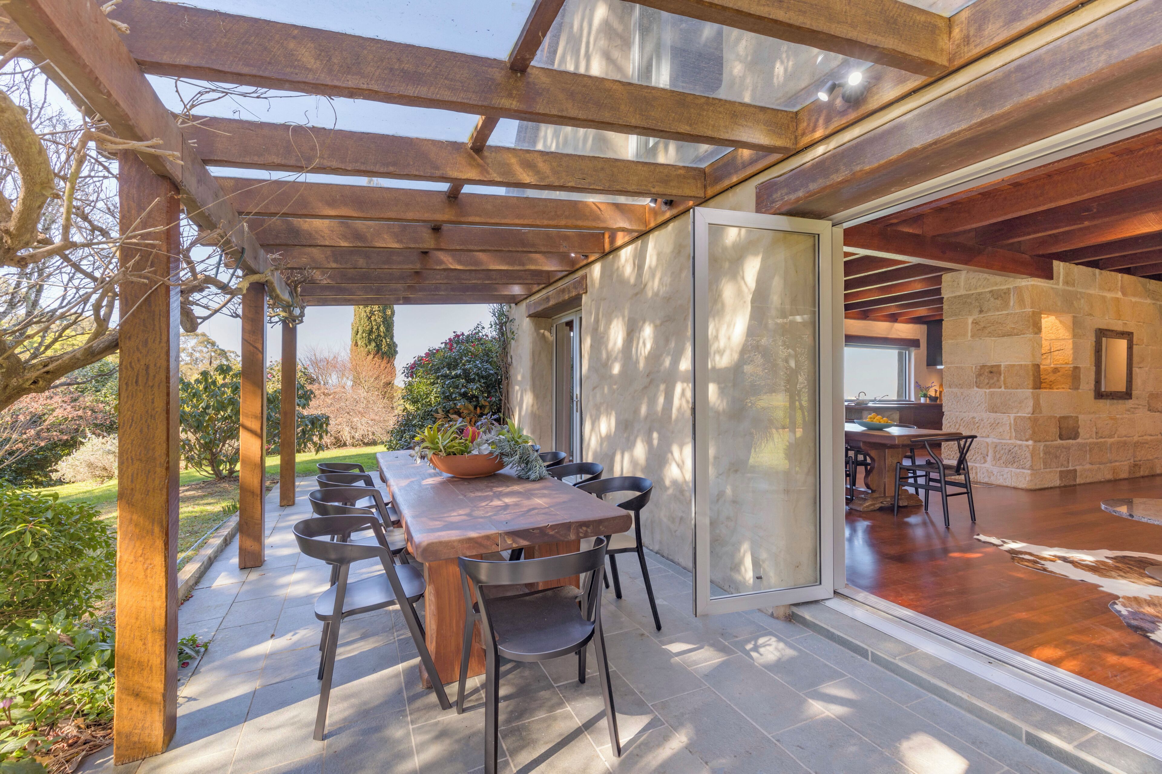 A covered patio with a wooden table and chairs, a stone wall, and a glass door leading to an interior space.