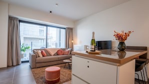 A modern living room with a sofa, ottoman, and a small coffee table. There is a kitchen area with a counter and a bottle of champagne. A large window provides a view of the city. - Baelskaai 25 with balcony by Interhome (Oostende)