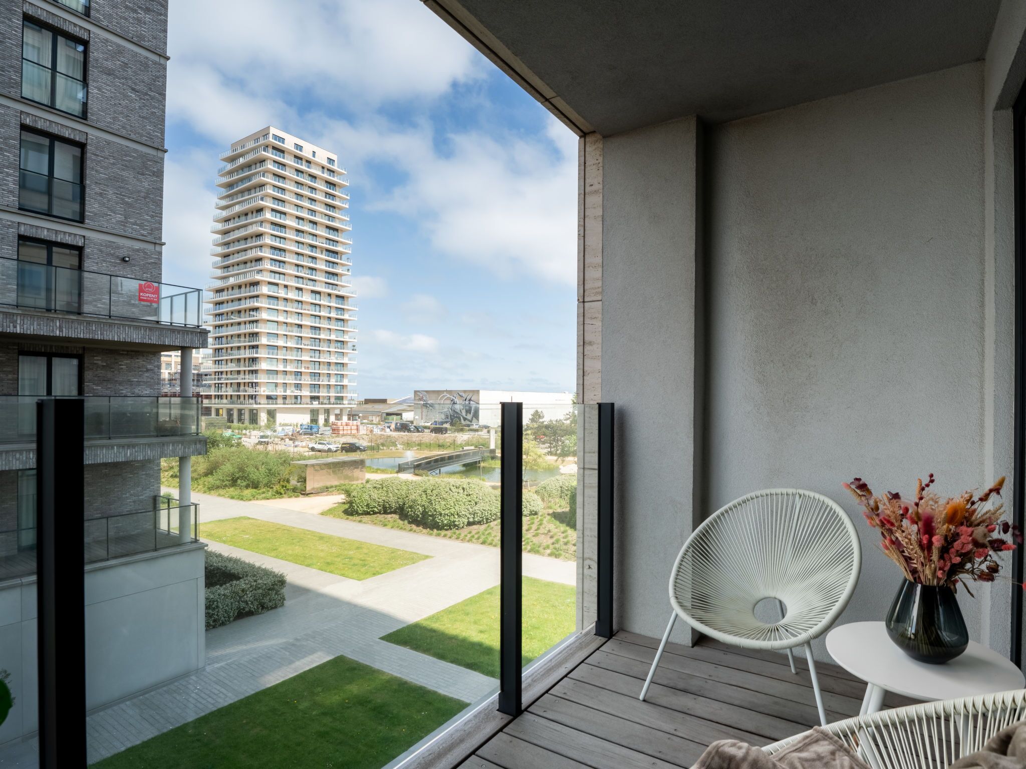 A balcony with a view of a high-rise building, a small table with a vase, and a white chair.
