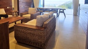 A modern living room with a large sofa, a wooden coffee table, and a dining area with a table and chairs. There is a ceiling fan and a large window providing a view of the outdoors. - Luxury Condo ALAMAR, Riviera Nayarit. Mexico (Cruz de Huanacaxtle)