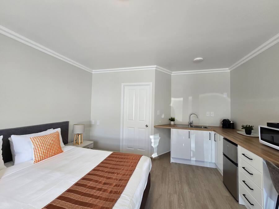 A modern bedroom with a bed, bedside table, lamp, and a kitchen area with cabinets and a sink.