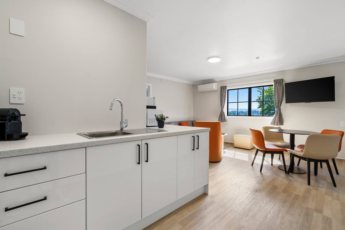 A modern kitchen with white cabinets, a sink, and a coffee maker. There is a dining area with a table and chairs, and a TV mounted on the wall.