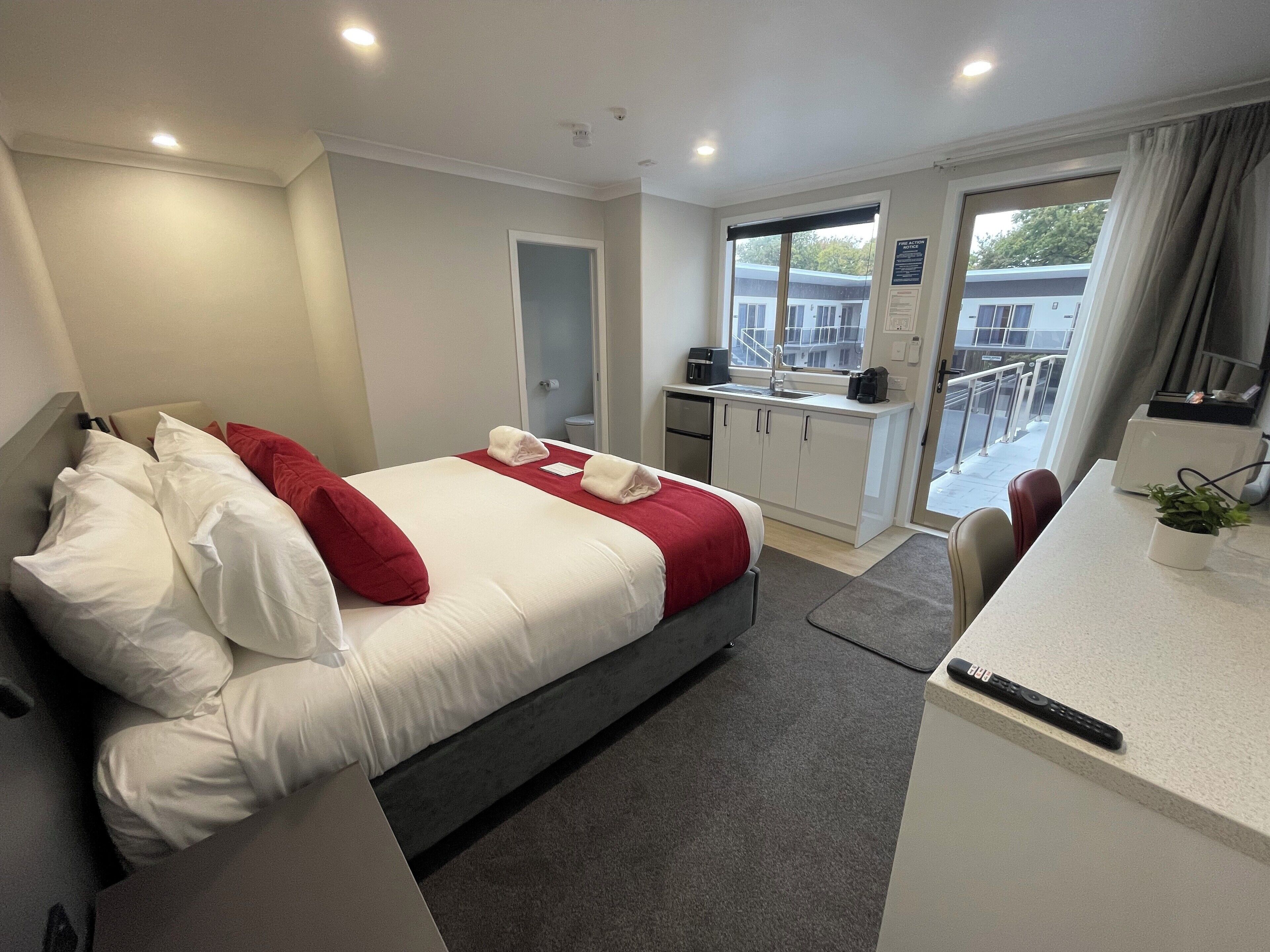A hotel room with a bed, a desk, a kitchenette, and a balcony with a view of buildings.
