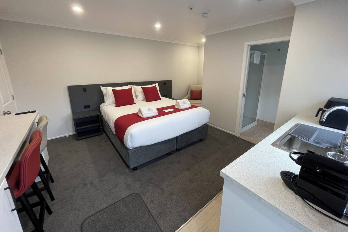 A modern hotel room with a bed, a desk, a chair, and a kitchenette.