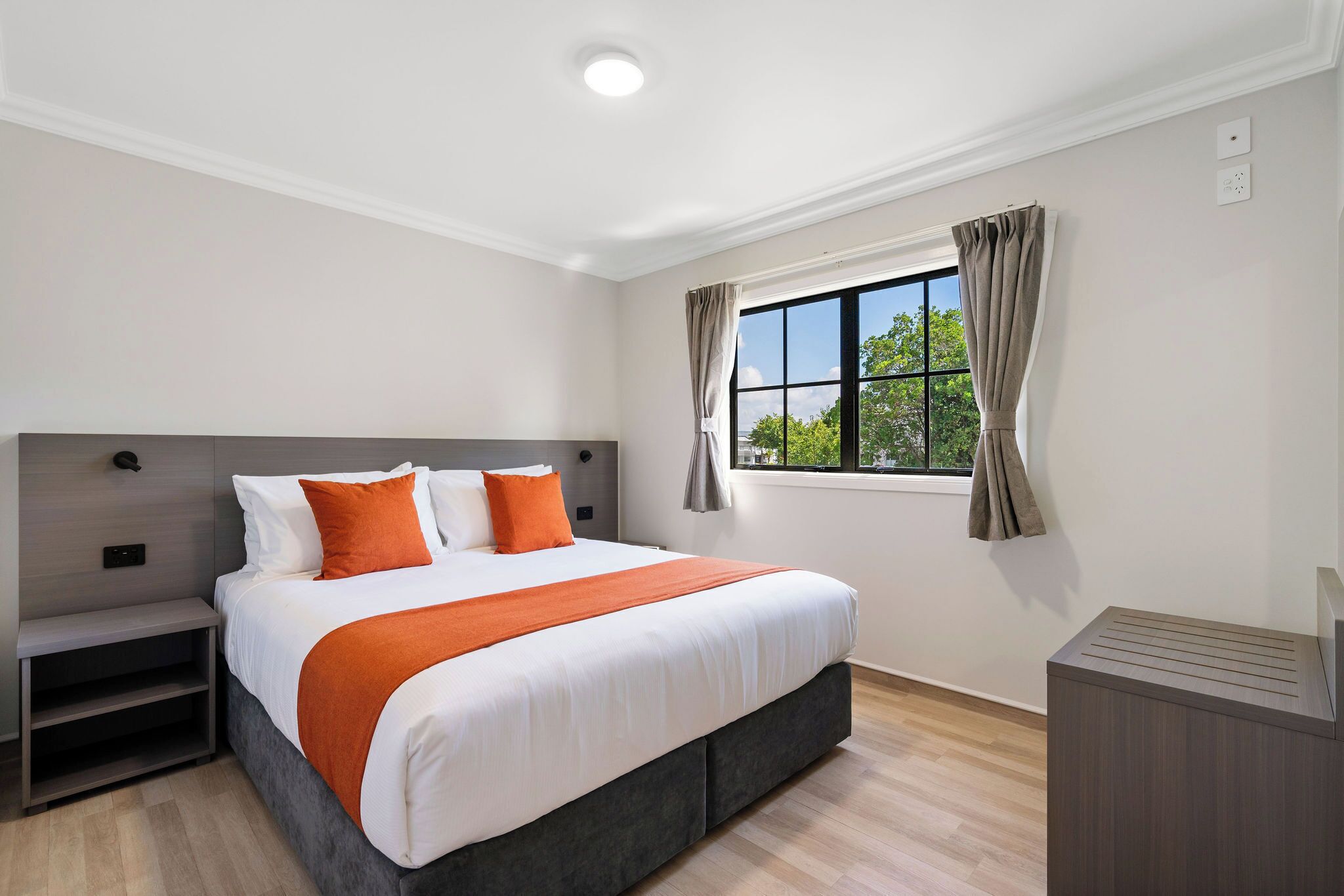 A modern hotel room with a large bed, a bedside table, a built-in heater, and a window with curtains.