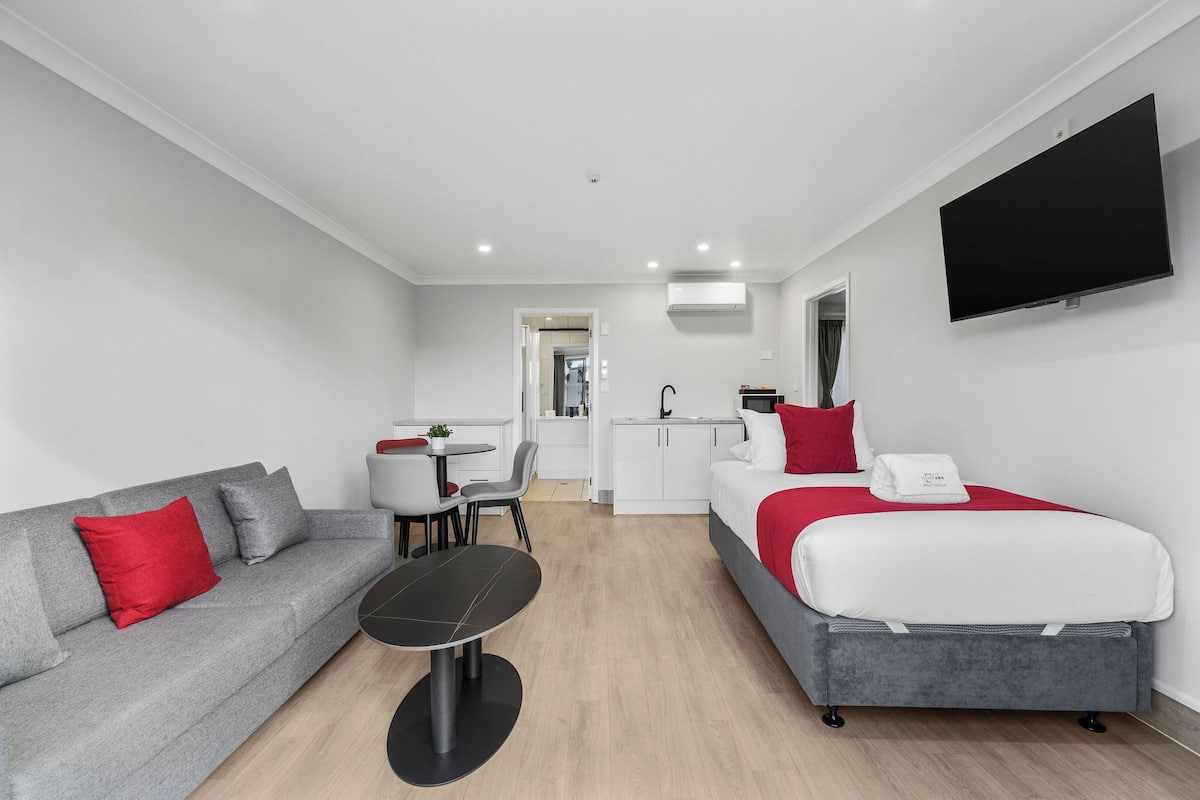A modern hotel room with a grey sofa, a small round table, a bed with red and white bedding, and a flat-screen TV mounted on the wall.