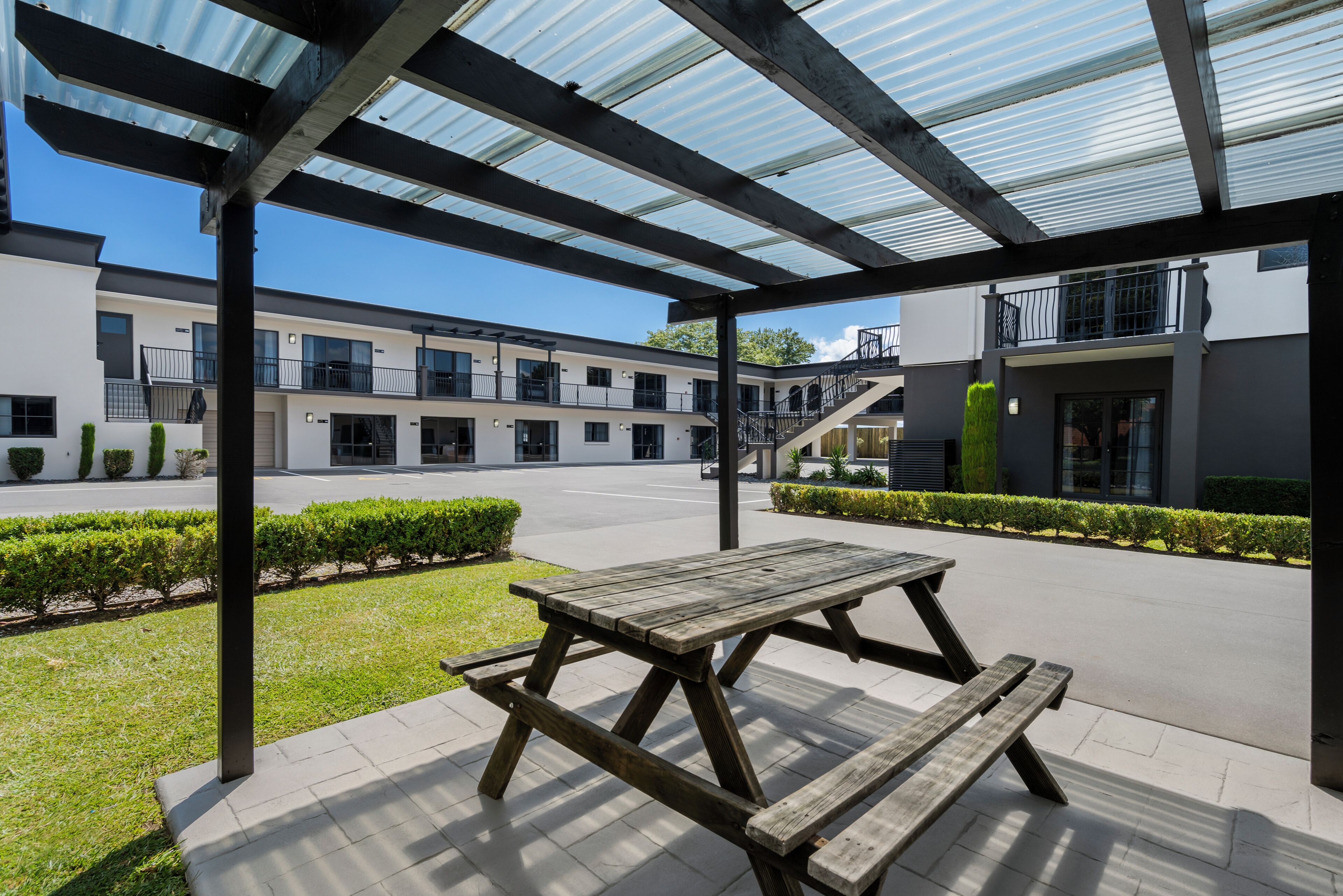 A covered outdoor area with a wooden picnic table, a clear sky, and a modern building with multiple windows and balconies in the background.
