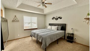 A bedroom with a bed, a ceiling fan, a window with blinds, a bedside table, and a wall-mounted decoration.