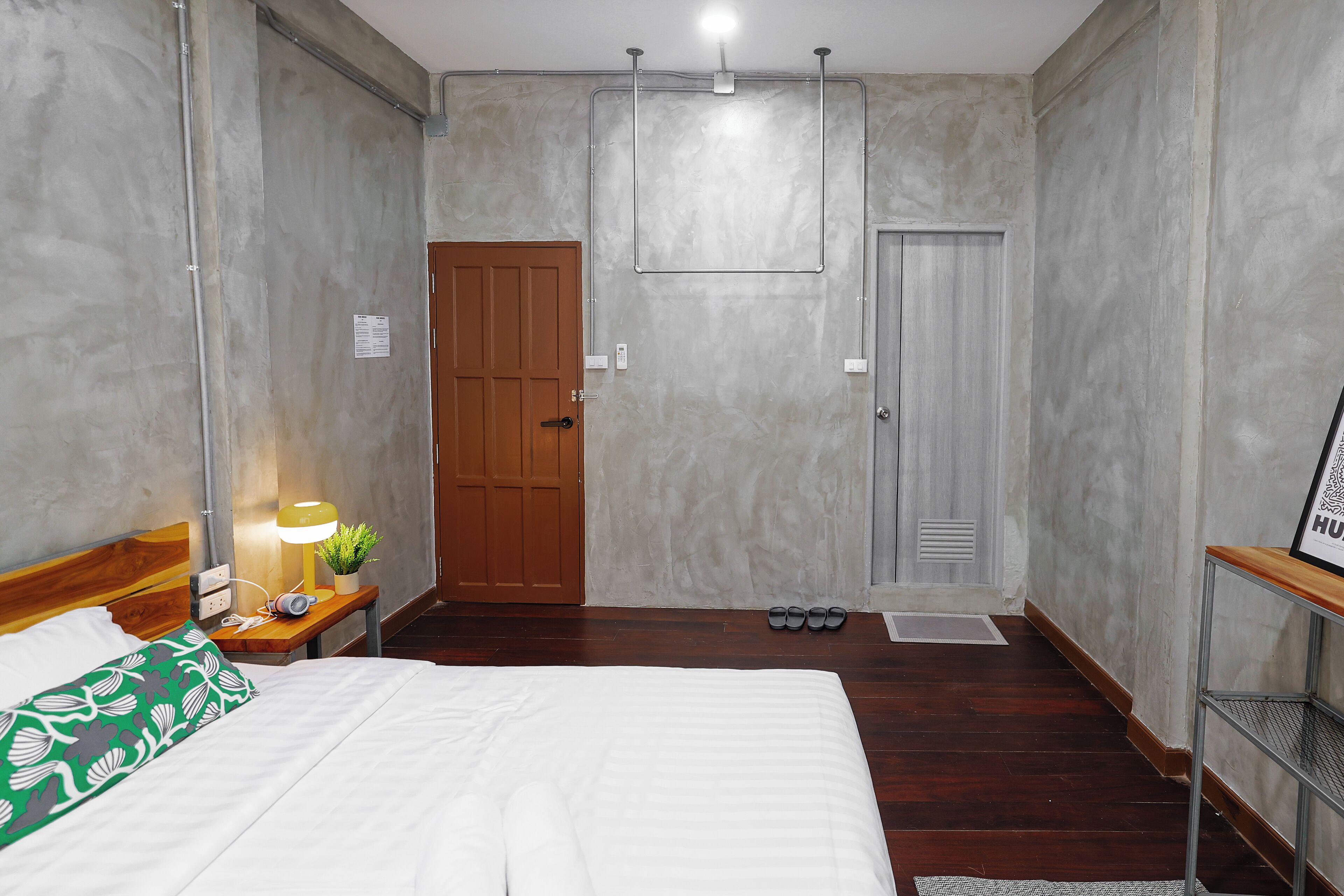 A hotel room with a bed, bedside table with a lamp, a small plant, and a framed picture on a shelf.