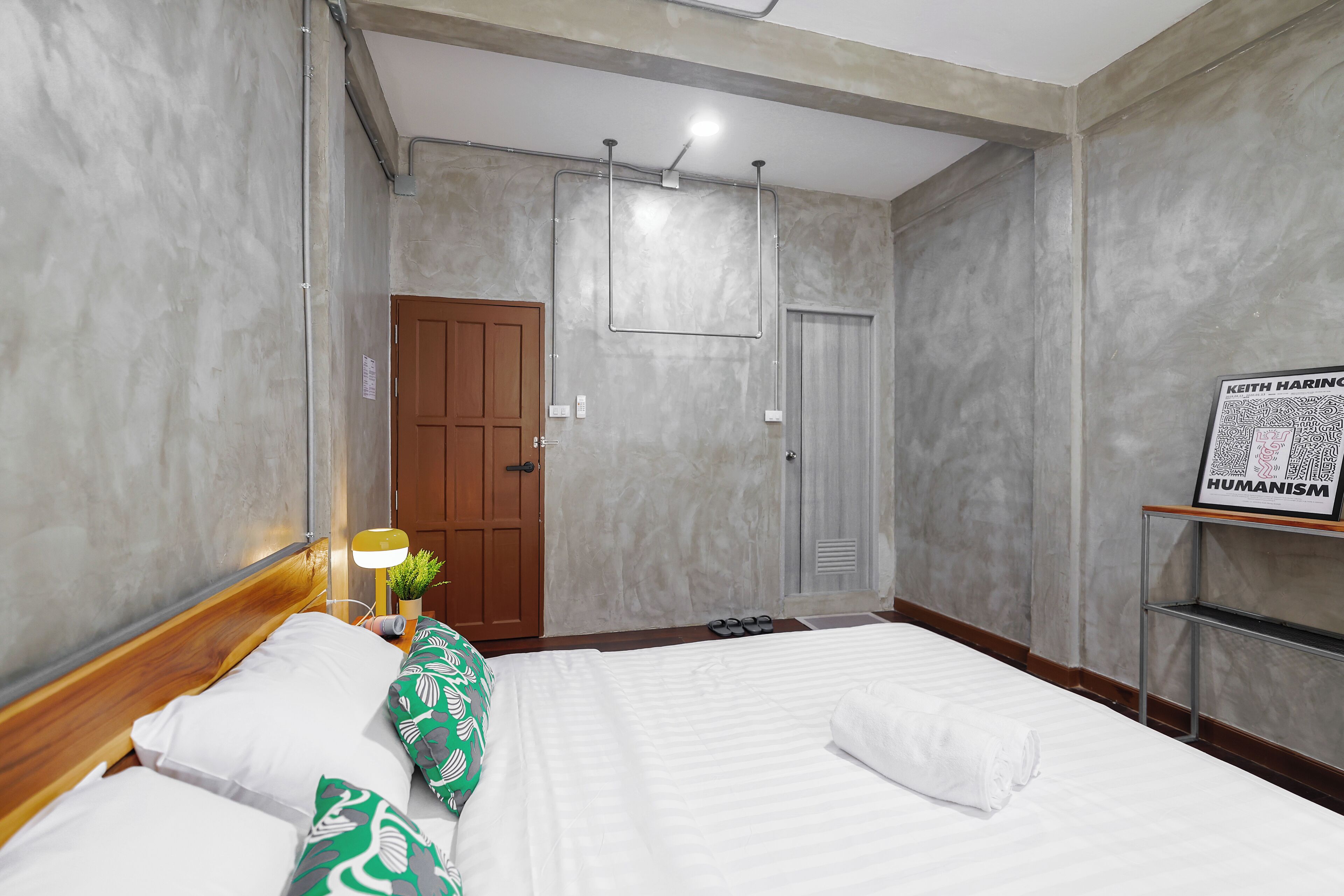 A hotel room with a wooden headboard, a bed with white linens, a green patterned pillow, a nightstand with a lamp, and a framed picture on the wall.