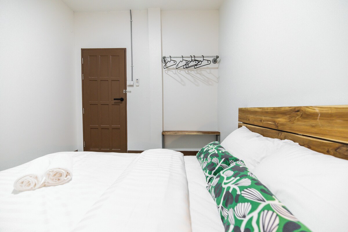 A bedroom with a wooden headboard, a bed with white bedding, a green floral-patterned pillow, and a coat rack with hangers.