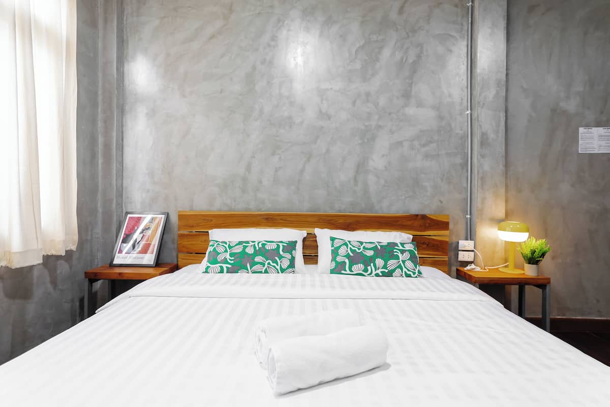 A neatly made bed with white bedding, two green patterned pillows, a folded white towel, a wooden headboard, a bedside table with a lamp and a framed picture, and a grey textured wall.