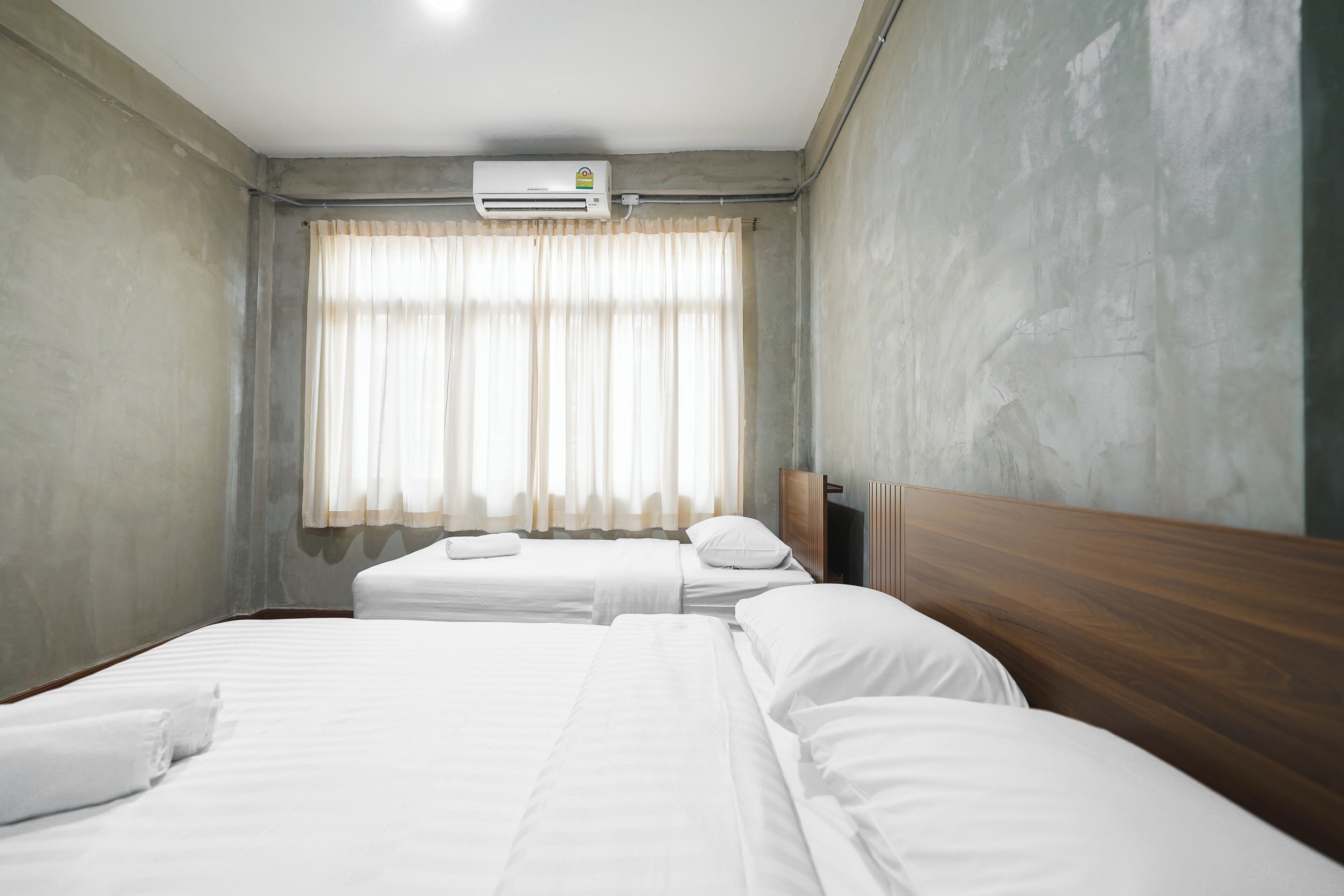 A hotel room with two beds, a window with curtains, an air conditioning unit, and a wooden headboard.