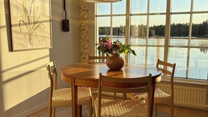 A dining area with a wooden table, woven chairs, a rug, a hanging light fixture, a framed botanical print, and a window with a view of a lake. - Lake House Retreat with a Dock & Boat (Västra Götalands län)