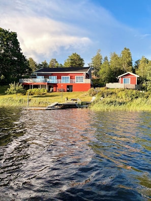Une maison rouge avec un quai qui s’avance dans l’eau, entourée de verdure et d’arbres.