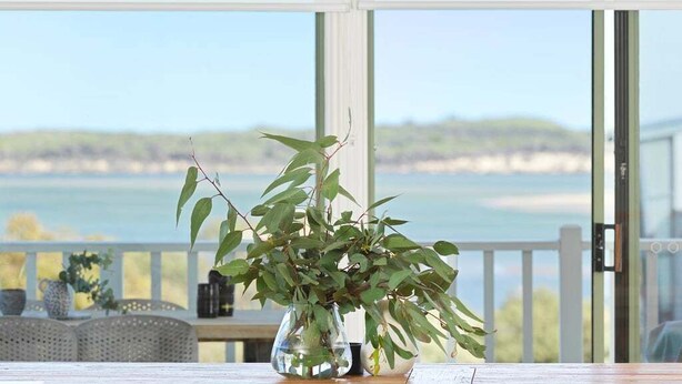 Ein Holztisch mit einer klaren Glasvase, gefüllt mit Grünpflanzen, befindet sich auf einem Balkon mit Blick auf ein Gewässer und das entfernte Land.