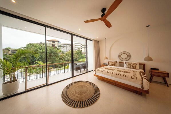 Une chambre moderne avec un grand lit, un ventilateur de plafond, une vue sur la verdure et un balcon.