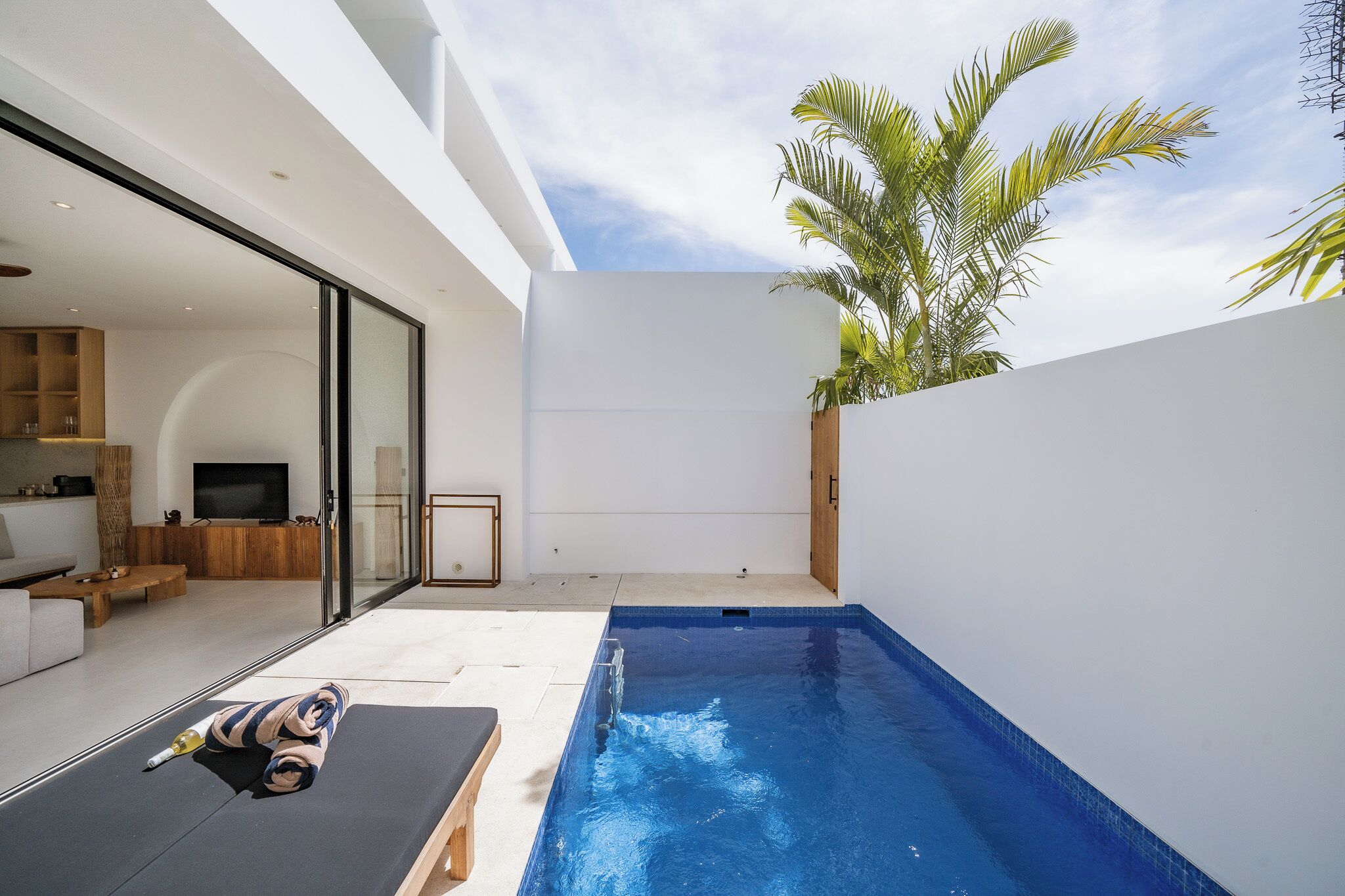 Un área de piscina exterior moderna con pared de vidrio, una silla de descanso y un televisor al fondo.