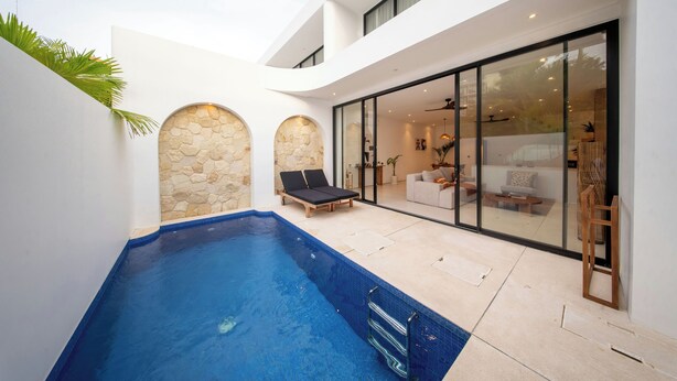 Un espace piscine extérieur moderne, agrémenté d’un mur en pierre, de deux chaises longues et d’une porte vitrée donnant sur un salon.
