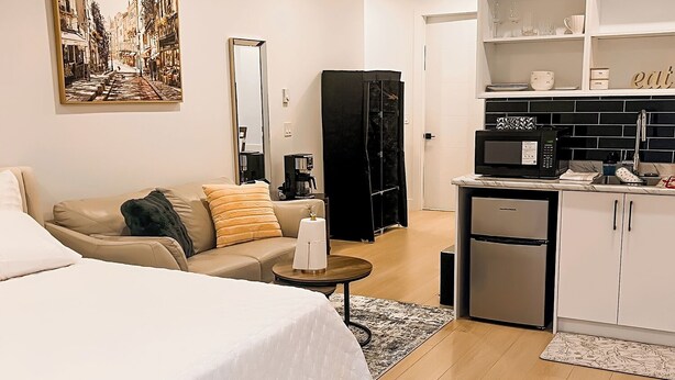 A modern hotel room with a large bed, a sofa, a coffee table, a black wardrobe, a kitchenette, and a dining area. - Tastefully furnished new studio in Bear Mountain (Langford)