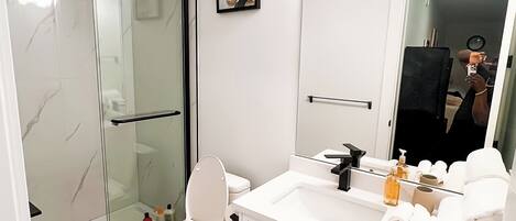 Ein modernes Badezimmer mit Duschkabine aus Glas, Toilette, Waschbecken mit Pflegeprodukten und Spiegel.