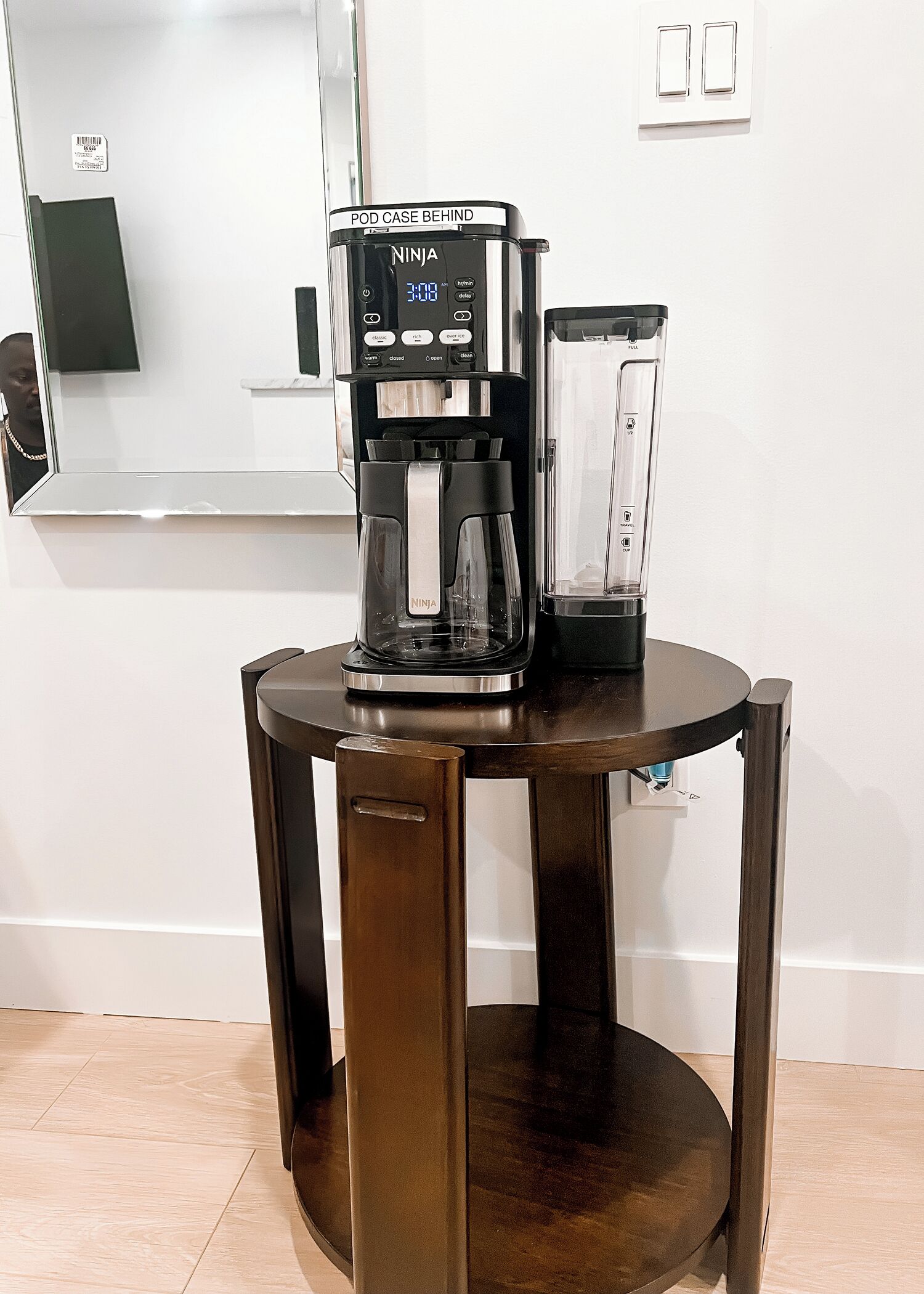 Une machine à café est posée sur une table ronde en bois. Un miroir et un interrupteur sont fixés au mur derrière la table.