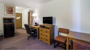 Habitación de hotel con escritorio, silla, televisor, microondas, refrigerador y una pequeña mesa con mantel estampado.