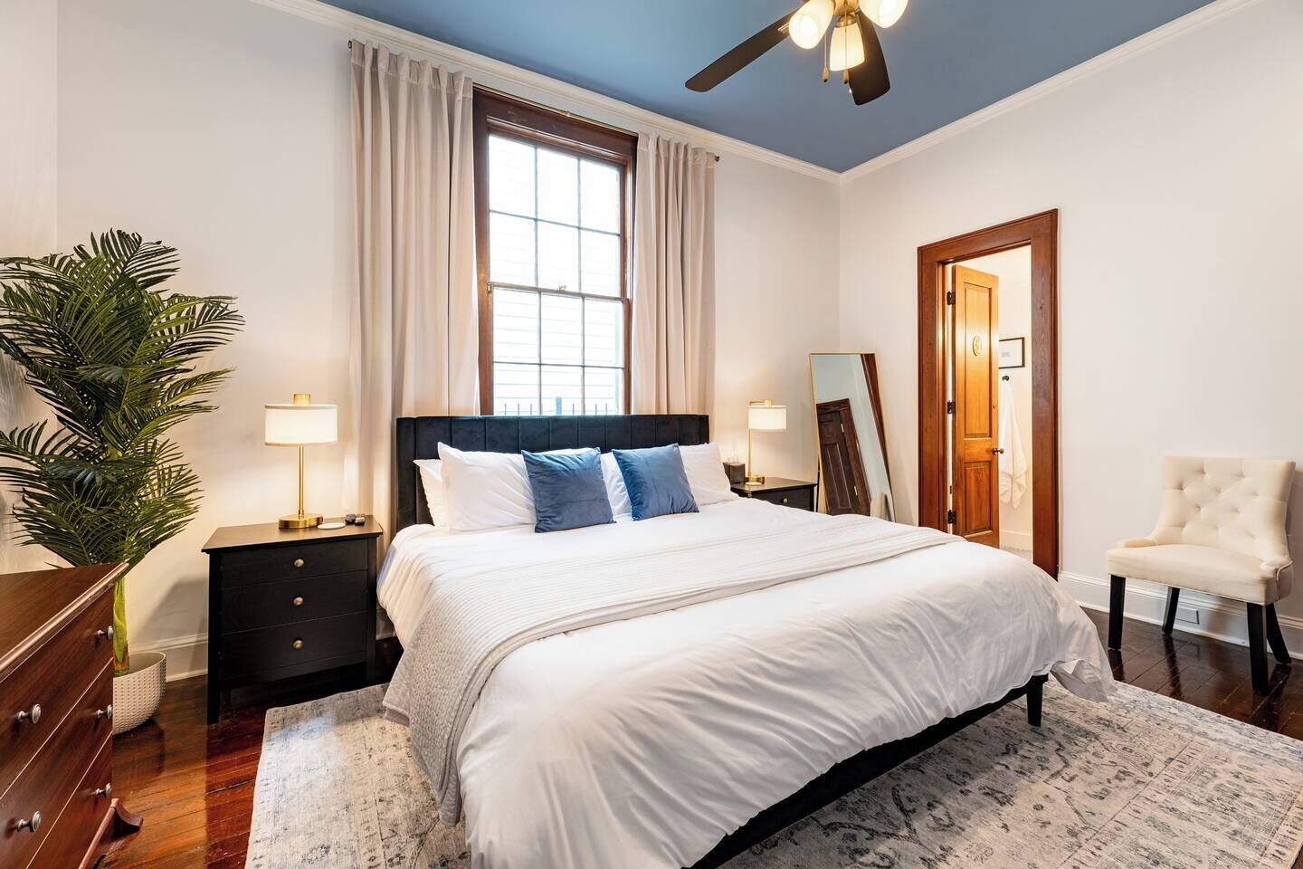 Une chambre avec un grand lit, un ventilateur de plafond, une fenêtre avec des rideaux, une table de chevet, une lampe, un miroir, une chaise et une plante.