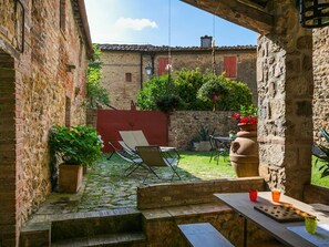 Une cour bordée de murs en pierre, une porte rouge, des sièges en plein air et des plantes en pot.