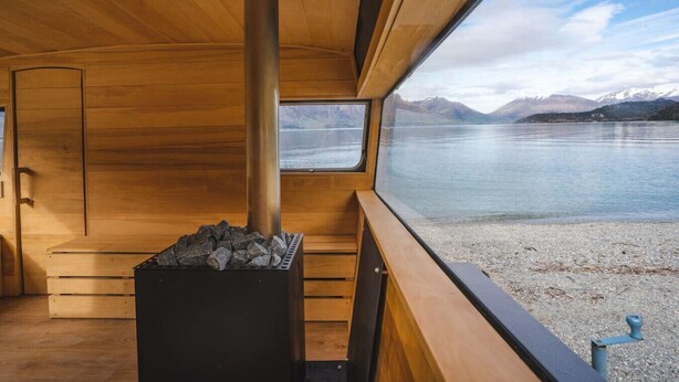 A cabin with a wood-burning stove, wooden walls, and a window overlooking a lake with mountains.