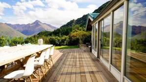 A wooden deck with a table and chairs, large glass windows, and a mountainous landscape view.