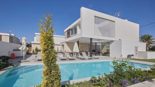 A modern two-story building with a swimming pool, lounge chairs, and a tall coniferous tree.