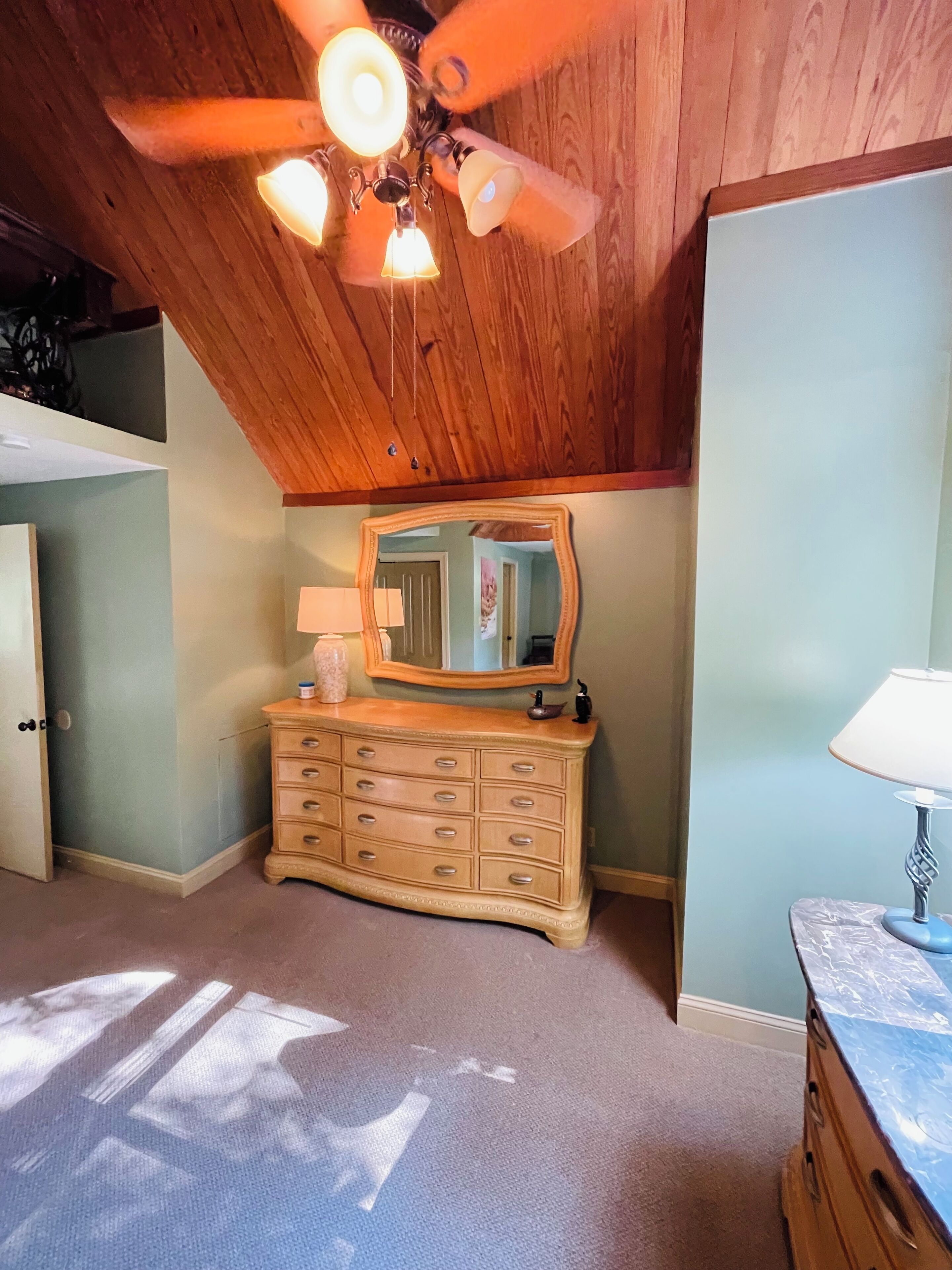 A room with a wooden ceiling, a dresser with drawers, a mirror, and a lamp.