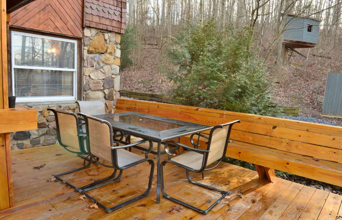 Eine Holzterrasse mit einem Tisch aus Glasplatte und Metallstühlen, umgeben von Steinmauern und einem Holzzaun.