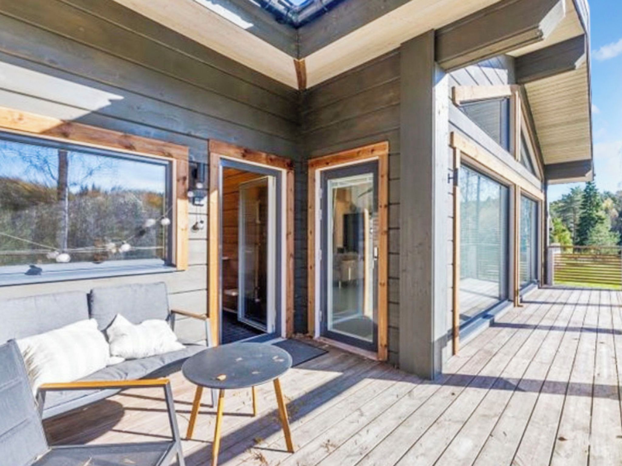 Une terrasse en bois avec un canapé et une table, de grandes portes vitrées donnant sur une pièce, et une vue sur les arbres.