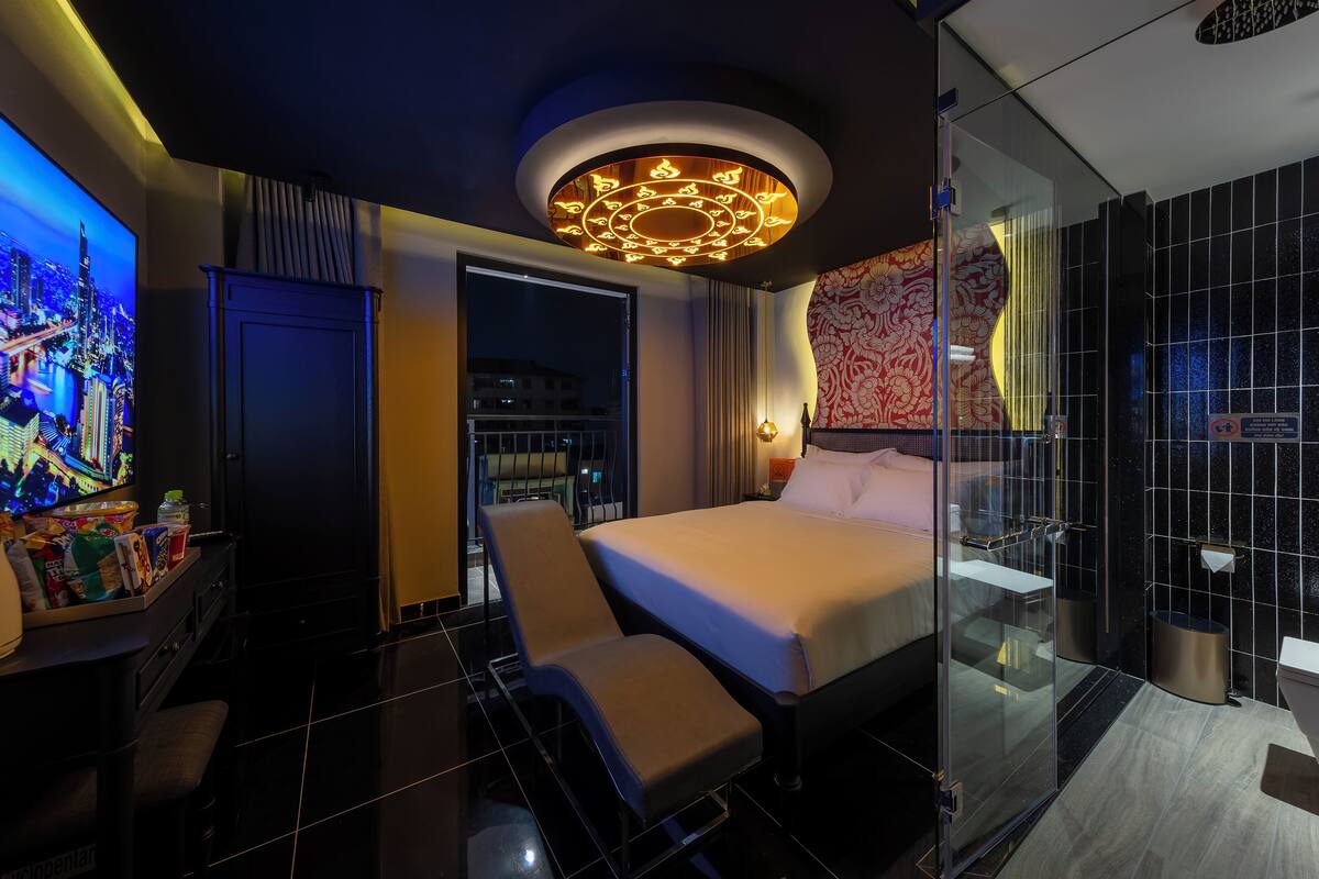 A modern hotel room with a large bed, a glass shower enclosure, and a cityscape view on the TV.
