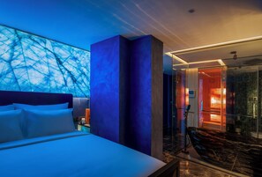 A modern hotel room with a large bed, a glass shower enclosure, and a view of the sky through the window.