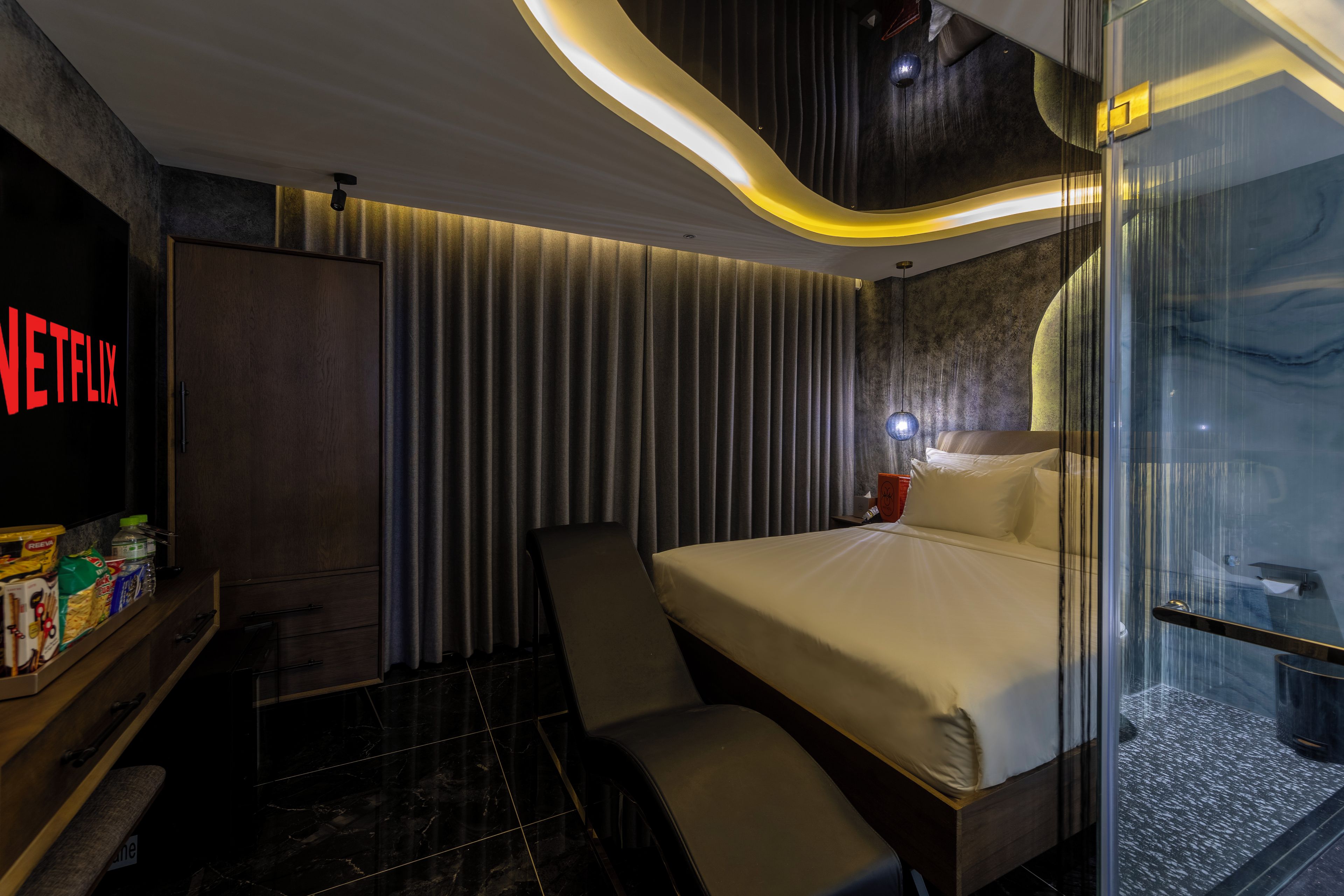 Una habitación de hotel moderna con una cama, zona de estar, minibar y mampara de vidrio.