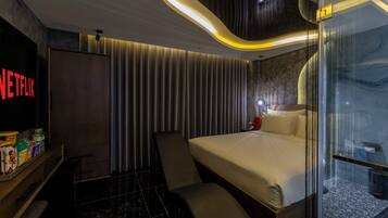Una habitación de hotel moderna con una cama, zona de estar, minibar y mampara de vidrio.
