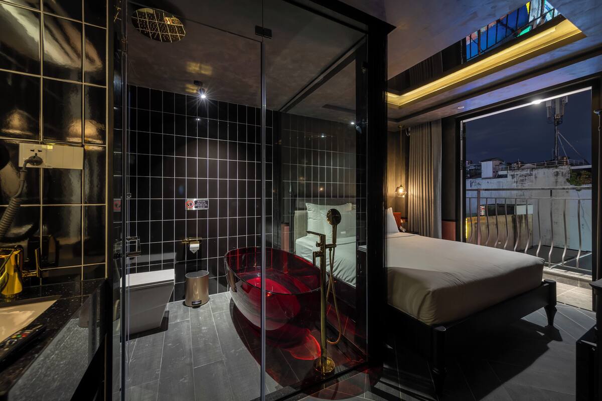 A modern bathroom with a glass-enclosed bathtub, a separate shower area, and a visible bed with a view of the cityscape.