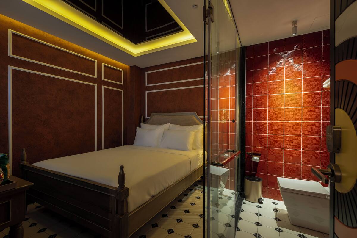 A modern hotel room with a large bed, a glass shower enclosure, and a red-tiled wall.