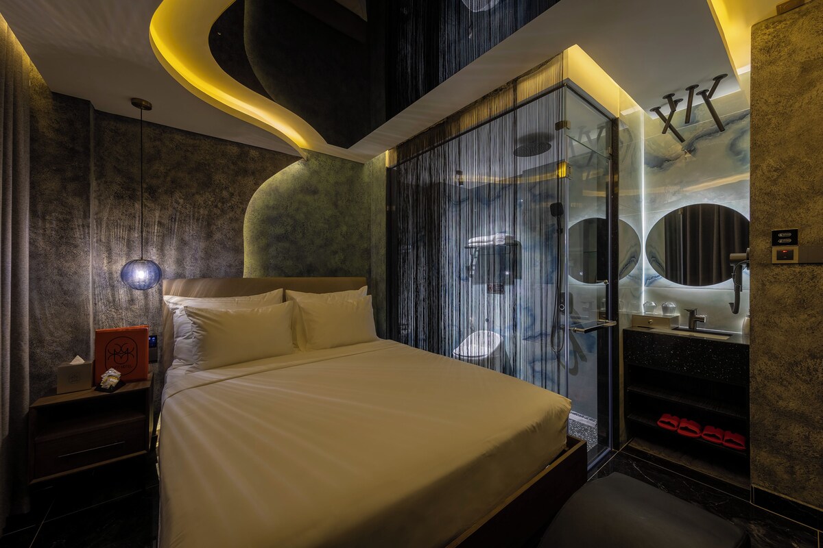 A modern hotel room with a large bed, a glass shower enclosure, and a vanity with a mirror.
