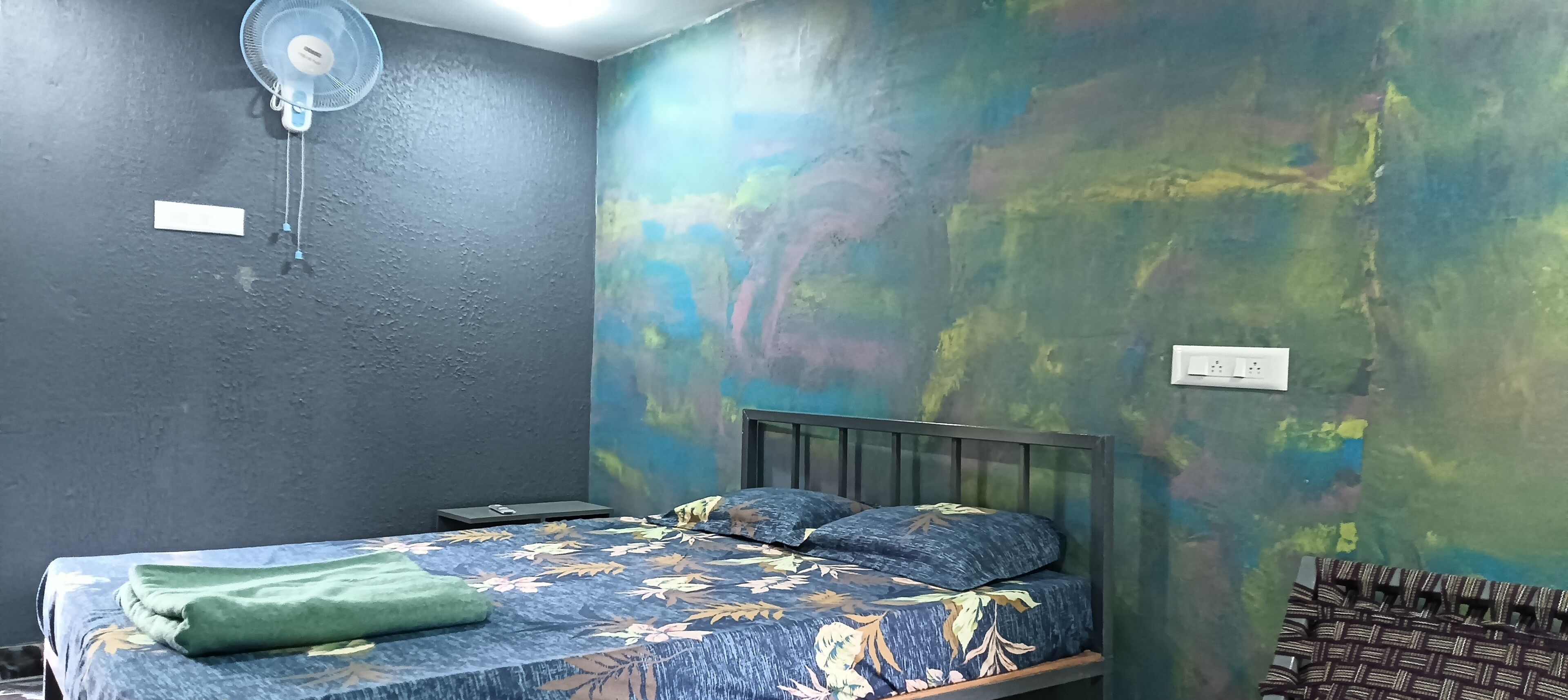 A room with a bed, a wall-mounted fan, and a colorful abstract wall mural.