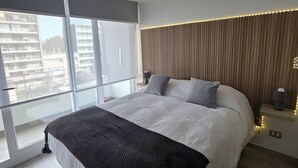 Ein modernes Schlafzimmer mit einem großen Bett, Blick auf Gebäude durch das Fenster und einem Nachttisch mit einer Lampe.