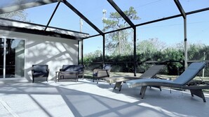 A covered outdoor seating area with chairs and a lounge chair, a small table, and a building with a white exterior. - This is it: Red Door Vacay House in Delightful Brandon with Game Room and Pool (Brandon)