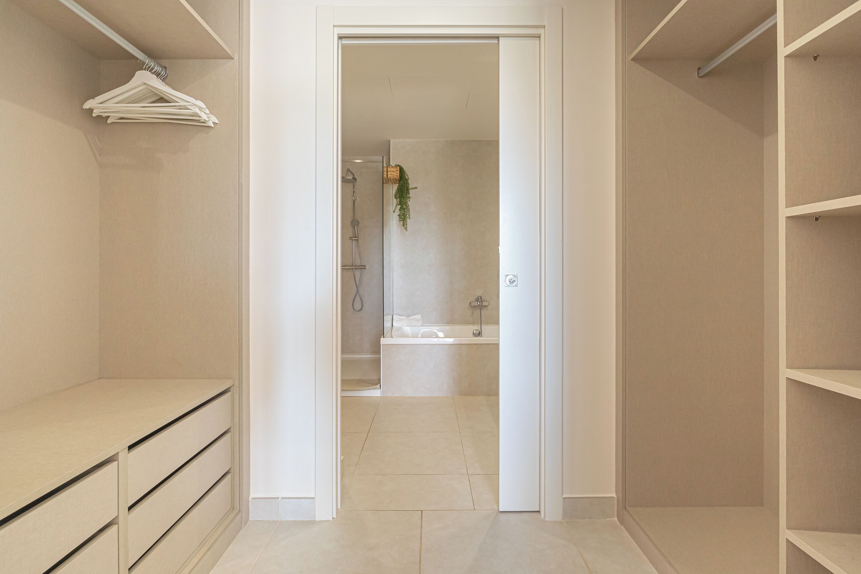 A walk-in closet with open doors leading to a bathroom with a shower, bathtub, and a hanging plant.