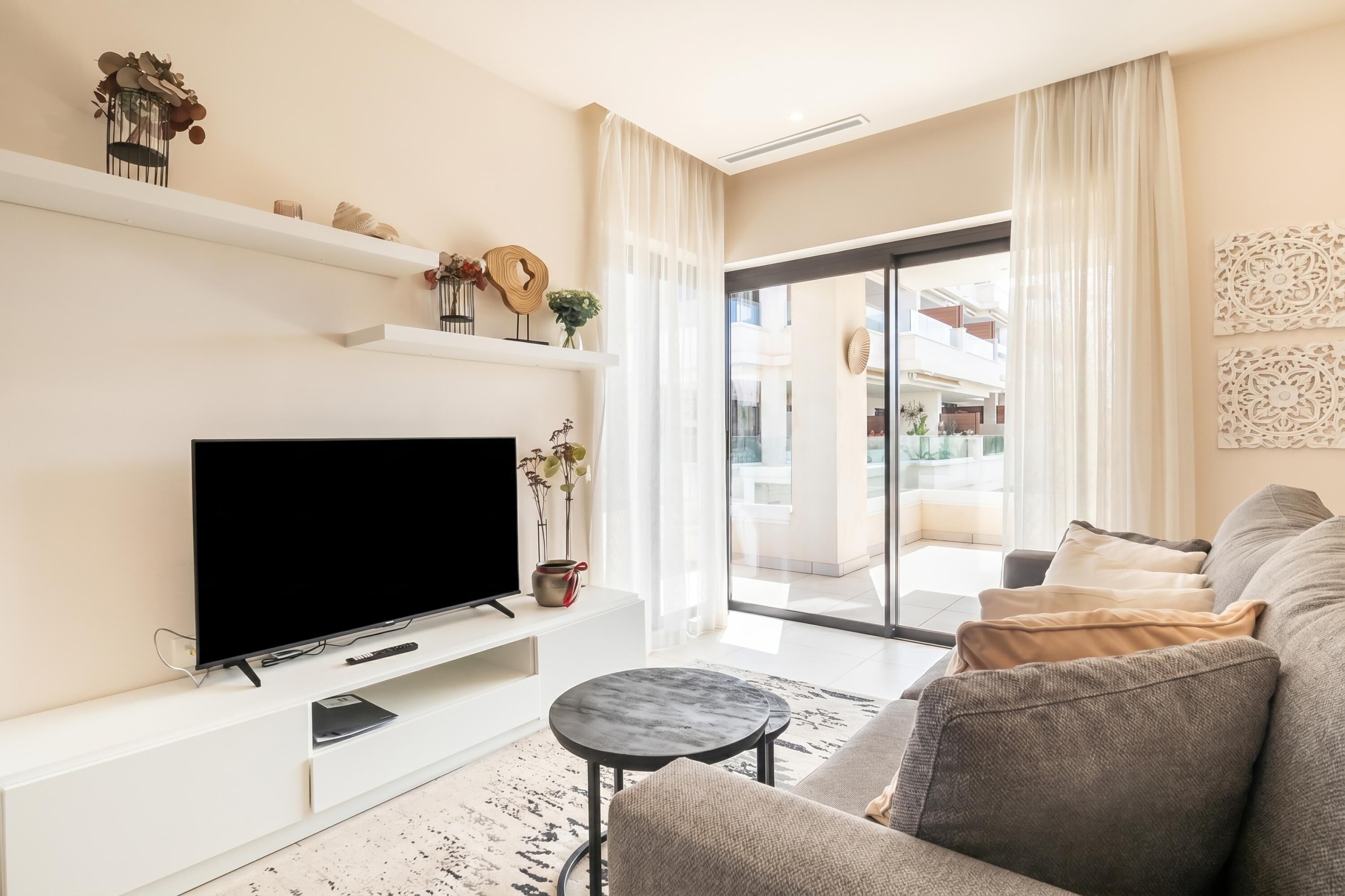 A modern living room with a flat-screen TV, a sofa, a coffee table, and a balcony with a view.