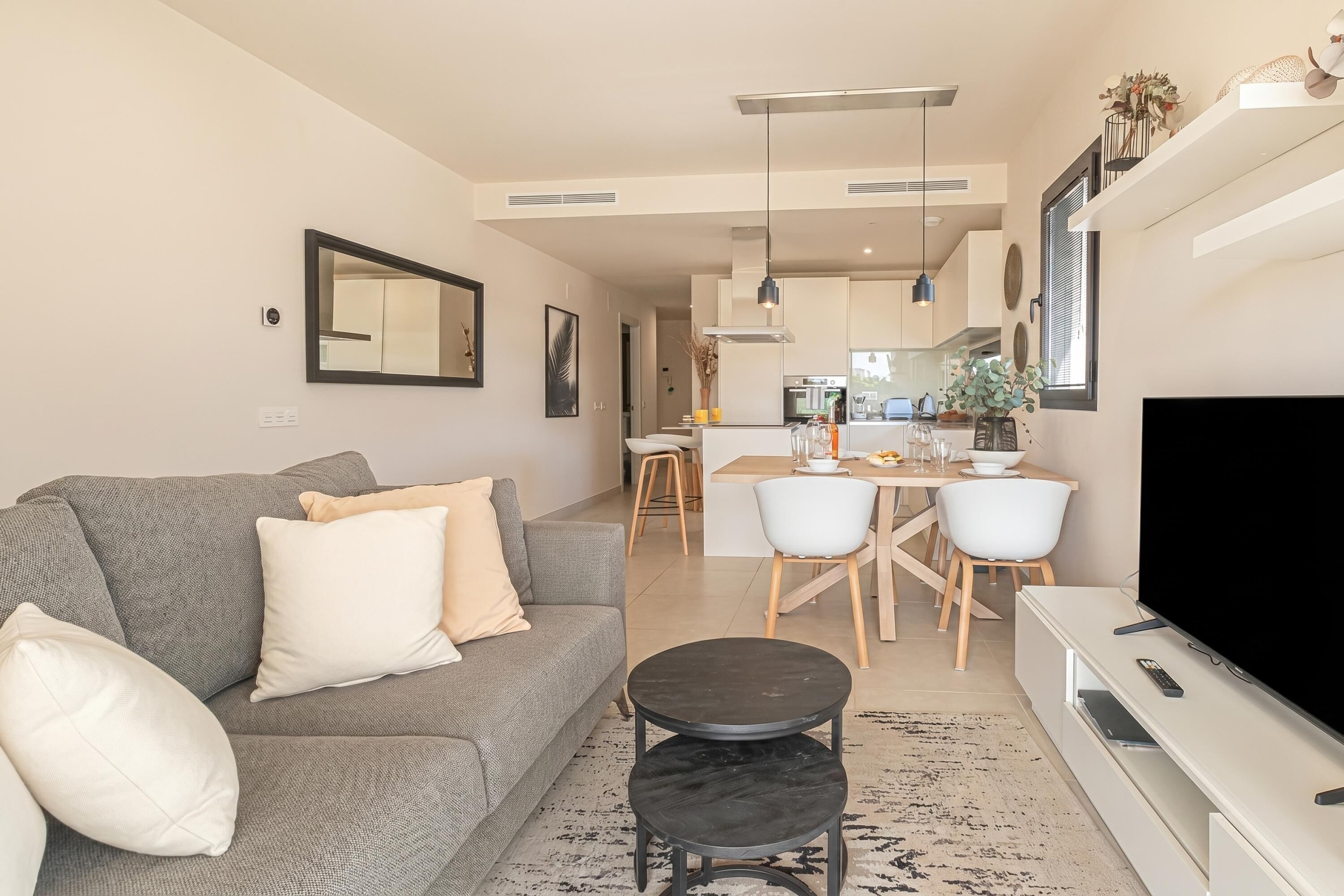 Una sala de estar moderna con un sofá gris, una mesa de centro, un televisor de pantalla plana y un comedor con una cocina al fondo.
