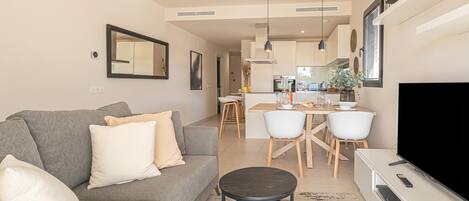 A modern living room with a grey sofa, a coffee table, a flat-screen TV, and a dining area with a kitchen in the background.